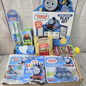 May include: A gift box containing Thomas & Friends merchandise. The contents include a play set, crayons, mini train toys, apple juice, snacks, and toothbrushes. The box features the blue and red Thomas the Tank Engine character.