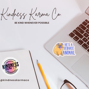 May include: A white laptop with a sticker of a cartoon pug with the text "He's a service animal" on the top right corner. A yellow pencil and a pair of glasses are on the table. A round sticker with the text "Kindness Karma Co." is on a notebook.