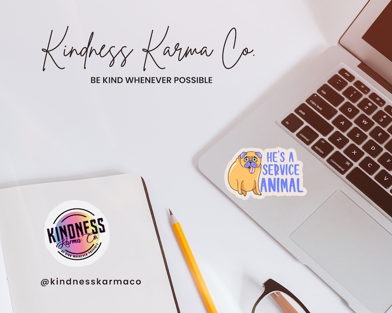 May include: A white laptop with a sticker of a cartoon pug with the text "He's a service animal" on the top right corner. A yellow pencil and a pair of glasses are on the table. A round sticker with the text "Kindness Karma Co." is on a notebook.