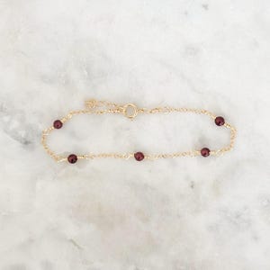 May include: A delicate gold bracelet with small, round, dark red beads. The bracelet has a simple chain design with the beads evenly spaced. The clasp is visible, and the bracelet is displayed on a white marble surface.