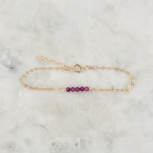 May include: A delicate gold-coloured bracelet featuring a series of small, round, deep pink gemstones. The bracelet has a simple chain design with a clasp closure. The gemstones are evenly spaced along the centre of the bracelet.