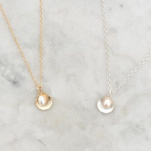 May include: Two seashell pendant necklaces, one gold and one silver. Each has a small pearl inside the shell. The chains are fine and the background is a marble effect. The necklaces are delicate.