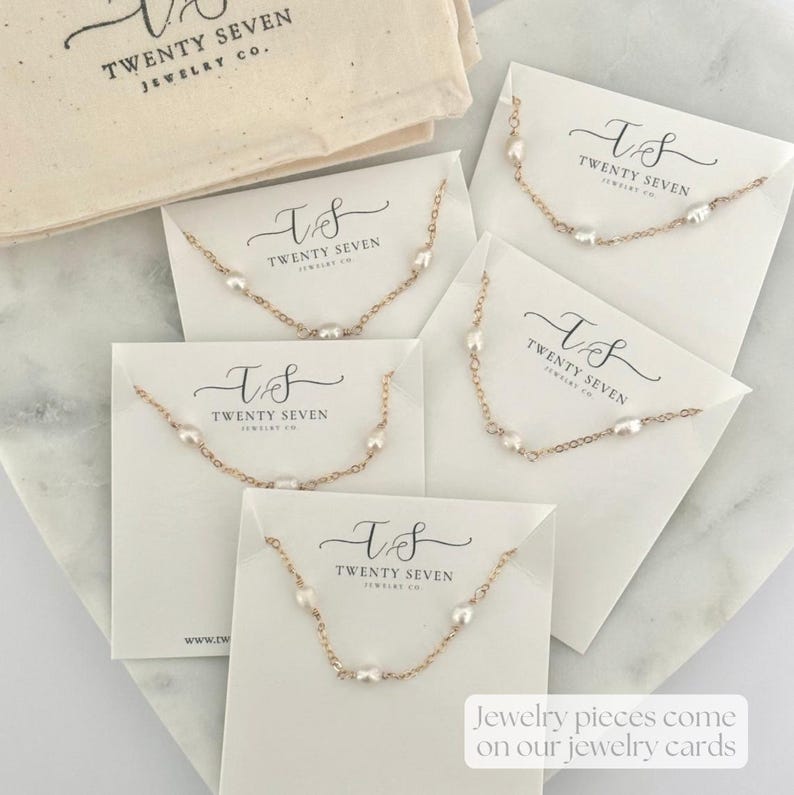 May include: Five white cards with gold chains and pearls on them. Each card has the text "T&S TWENTY SEVEN JEWELRY CO." printed on it. The cards are on a white surface. Jewelry pieces come on our jewelry cards.
