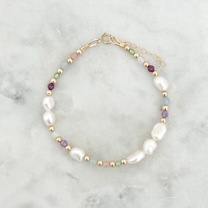 Könnte beinhalten: Ein zartes Armband mit weißen Süßwasserperlen, kleinen goldenen Perlen und farbigen Edelsteinen. Die Edelsteine umfassen rosa, grüne, violette und blaue Farbtöne. Das Armband wird mit einem goldenen Verschluss gesichert.