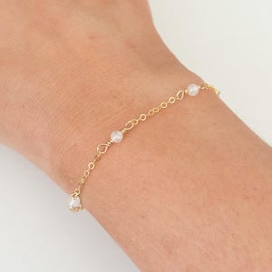 May include: A delicate gold chain bracelet with small white beads. The bracelet is on a person's wrist.
