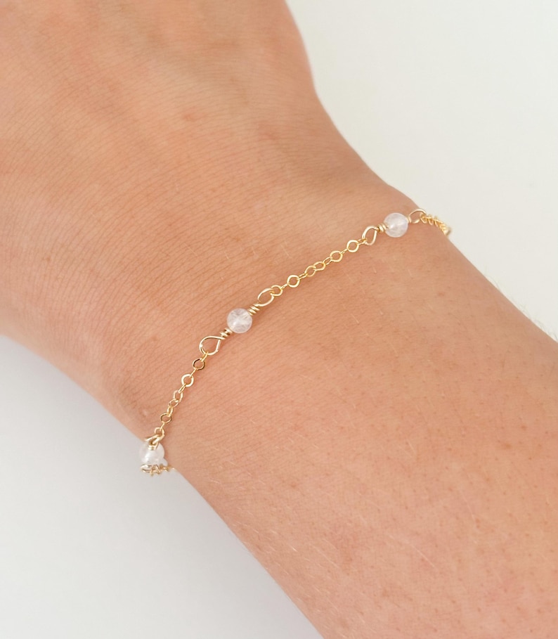 May include: A delicate gold chain bracelet with small white beads. The bracelet is on a person's wrist.