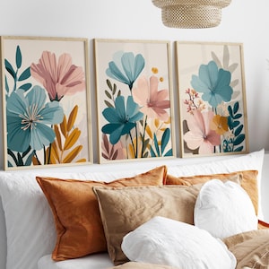 Puede incluir: Tres impresiones artísticas florales enmarcadas. Cada impresión presenta flores estilizadas en tonos turquesa, rosa y amarillo sobre un fondo neutro. Los marcos son de madera clara. Las impresiones se exhiben sobre una cama con almohadas naranjas y blancas.