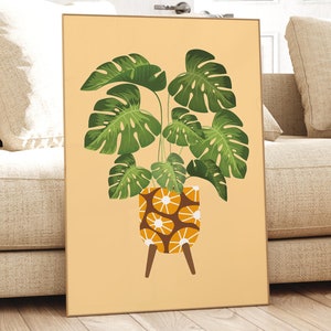 Puede incluir: Una impresión enmarcada de una planta de monstera verde en una maceta marrón y naranja con un patrón geométrico blanco. La maceta tiene cuatro patas de madera. El fondo es de color melocotón claro.