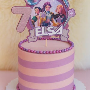 Peut inclure: Un gâteau cylindrique à rayures horizontales rose clair et violet, décoré de bonbons sphériques violets et roses sur les bords supérieur et inférieur. Une décoration de gâteau violette pailletée affiche le chiffre 7, le nom ELSA et trois personnages.