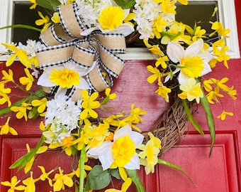 May include: A spring wreath with yellow daffodils, white flowers, and green leaves. A burlap bow with a black and white checkered pattern is centered on the wreath. The wreath is displayed on a red door.