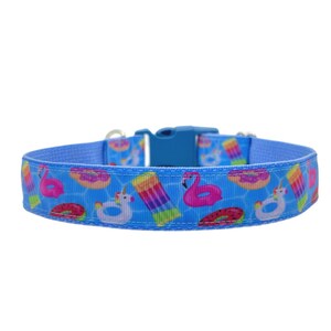 May include: A blue dog collar with a plastic buckle. The collar features a pattern of pool floats, including pink flamingos, donuts, unicorn floats, and rainbow popsicles, set against a blue water background.