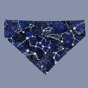 May include: A triangular pet bandana featuring a dark blue background with a constellation pattern in white. The design includes numerous white stars and lines, creating a celestial theme. The bandana is likely made of fabric.
