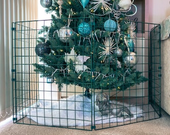 Op de afbeelding: Een groene metalen poort omringt een gedecoreerde kerstboom met blauwe en zilveren ornamenten. Een witte, harige boomrok bedekt de basis van de boom. Een klein grijs hondenbeeldje ligt op de boomrok.