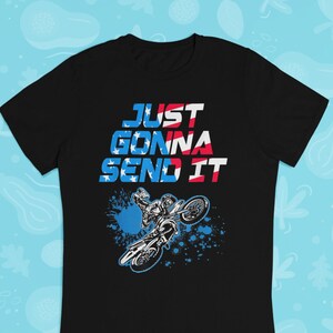 Puede incluir: Camiseta negra con un gr&aacute;fico de un piloto de motocross saltando sobre una rampa. El gr&aacute;fico es en azul, rojo y blanco, y el texto "JUST GONNA SEND IT" est&aacute; impreso sobre el gr&aacute;fico.