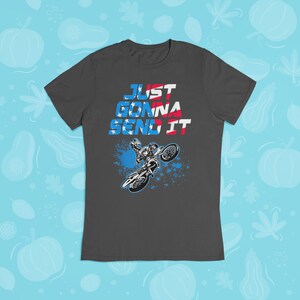 Puede incluir: Una camiseta gris oscuro con el texto "JUST GONNA SEND IT" en rojo, blanco y azul. Un gr&aacute;fico de un piloto de motocross realizando un truco est&aacute; debajo del texto.