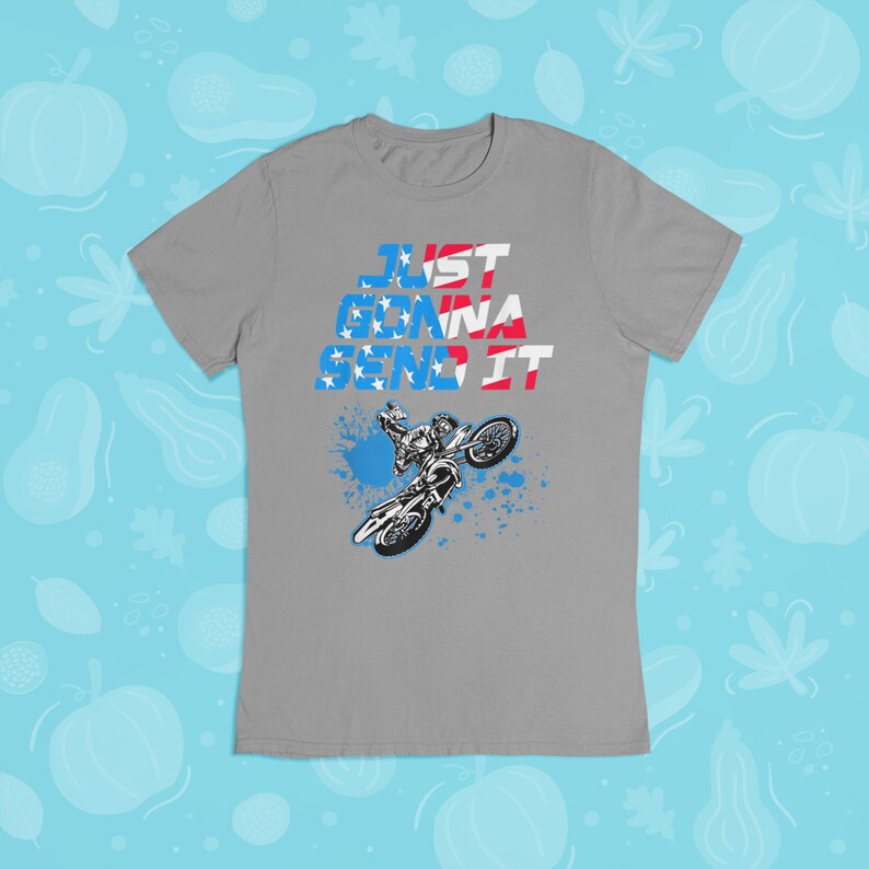 Puede incluir: Camiseta gris con un gr&aacute;fico de un piloto de motocross realizando un truco. El texto "JUST GONNA SEND IT" est&aacute; impreso en la camiseta en rojo, blanco y azul.