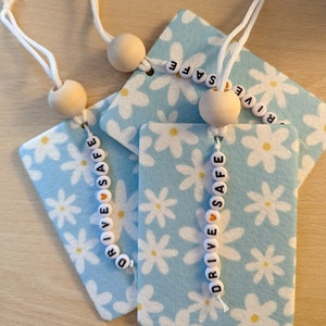 May include: Three blue and white car air fresheners with a daisy pattern. Each air freshener has a white beaded necklace with the words "DRIVE SAFE" spelled out in black letters.