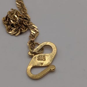 May include: Close-up of a gold-colored necklace with a textured chain and a hook clasp. The clasp is marked with the number "916". The necklace is likely made of gold and is designed to be worn around the neck.
