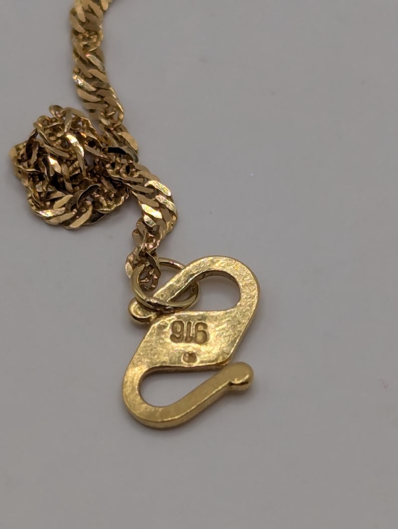May include: Close-up of a gold-colored necklace with a textured chain and a hook clasp. The clasp is marked with the number "916". The necklace is likely made of gold and is designed to be worn around the neck.