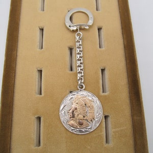 May include: A silver and gold keychain featuring a round pendant with an embossed image of a person wearing a headdress. The keychain has a silver chain and a silver ring.