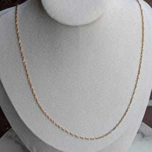 May include: A delicate gold-colored necklace with a twisted rope design. The necklace is displayed on a white mannequin bust against a neutral background. The chain is thin and appears to be made of metal.