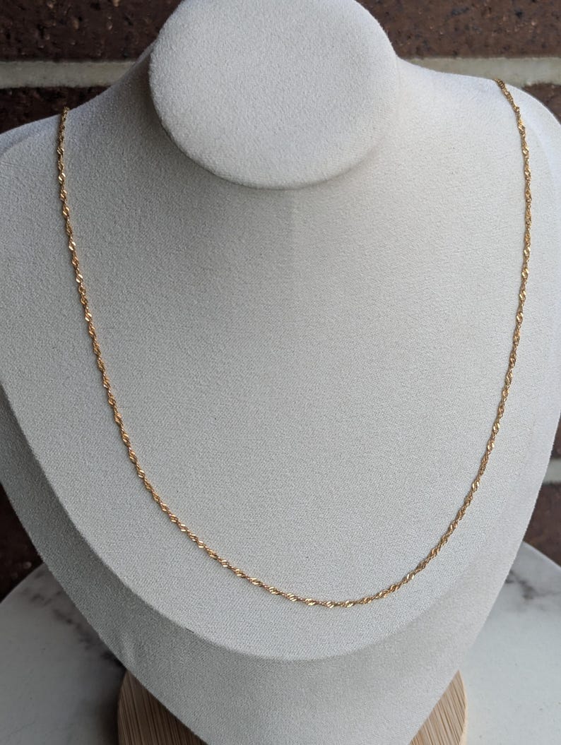 May include: A delicate gold-colored necklace with a twisted rope design. The necklace is displayed on a white mannequin bust against a neutral background. The chain is thin and appears to be made of metal.