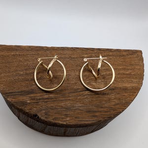 Puede incluir: Un par de pendientes de aro de oro con un diseño trenzado. Los pendientes están sobre una superficie de madera.
