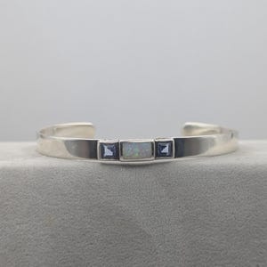 May include: A silver cuff bracelet with a rectangular opal center stone, flanked by two square blue stones. The bracelet has a simple, modern design.