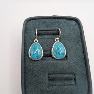 Pode incluir: Um par de brincos turquesa em forma de gota com molduras de metal prateado. Os brincos são exibidos numa caixa de joias verde escuro. As pedras turquesas têm uma aparência marmoreada com tons de azul e branco.