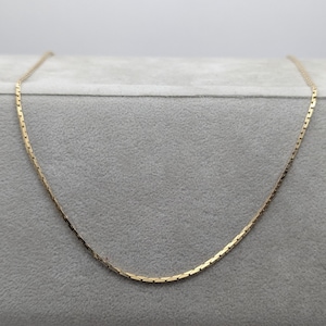 Könnte beinhalten: Eine zarte, goldfarbene Halskette mit einer flachen, rechteckigen Kettengliederkette. Die Kette wird auf einer grauen Oberfläche präsentiert, die ihr schlichtes, aber elegantes Design zeigt. Die Halskette ist ein klassisches Schmuckstück.