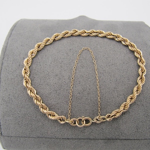 May include: A gold-colored rope chain bracelet with a safety chain. The bracelet is displayed on a gray surface. The bracelet has a clasp closure.
