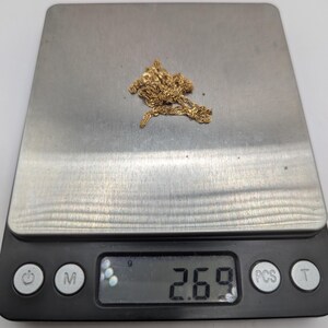 May include: A gold-colored chain necklace rests on a digital scale, displaying a weight of 2.69. The scale is black with silver-colored weighing surface. The necklace is a delicate design, and the background is white.