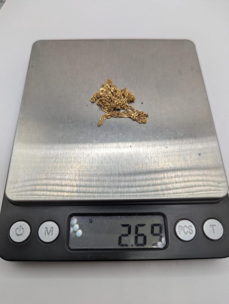 May include: A gold-colored chain necklace rests on a digital scale, displaying a weight of 2.69. The scale is black with silver-colored weighing surface. The necklace is a delicate design, and the background is white.