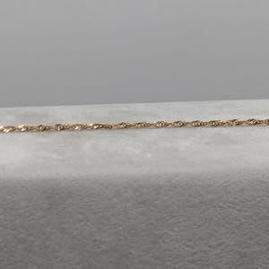 May include: A delicate gold-colored chain necklace with a twisted rope design. The necklace is displayed on a light gray surface, with a plain gray background. The chain is thin and appears to be made of metal.