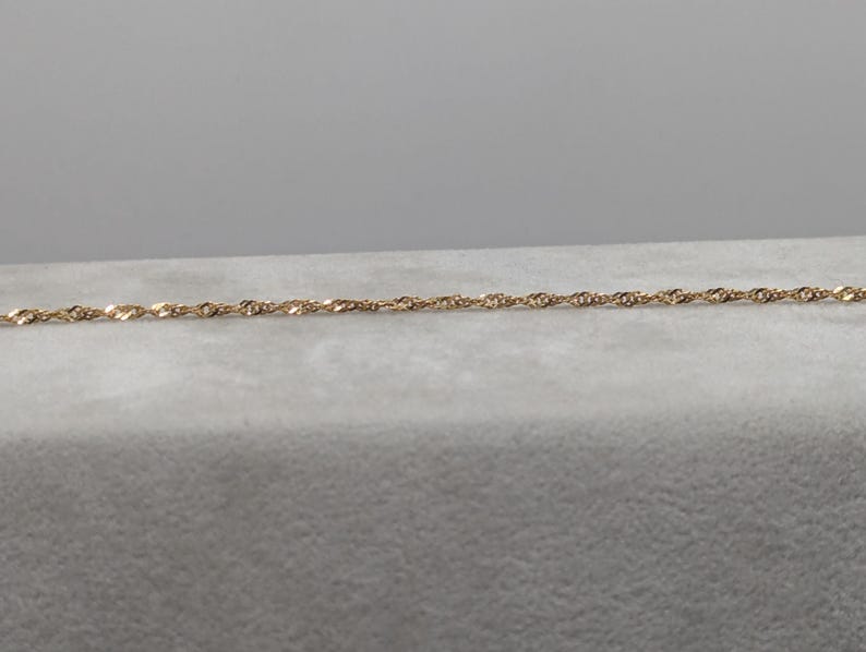 May include: A delicate gold-colored chain necklace with a twisted rope design. The necklace is displayed on a light gray surface, with a plain gray background. The chain is thin and appears to be made of metal.