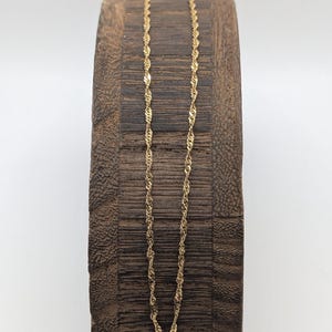 May include: A delicate gold-colored chain necklace with a twisted rope design. The necklace is displayed on a dark brown, textured wooden block against a white background. The chain's intricate pattern catches the light, showcasing its subtle elegance.