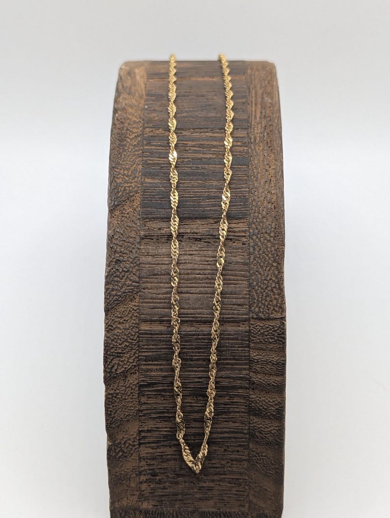 May include: A delicate gold-colored chain necklace with a twisted rope design. The necklace is displayed on a dark brown, textured wooden block against a white background. The chain's intricate pattern catches the light, showcasing its subtle elegance.