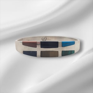 May include: A silver bracelet with a geometric design. The bracelet features rectangular inlays of various colours, including brown, black, blue, and green. The bracelet is set against a white background.