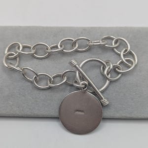 May include: A silver bracelet with a toggle clasp and a circular charm. The bracelet features a chain link design with a round charm that has the word "STERLING" stamped on it. The bracelet is displayed on a gray surface.