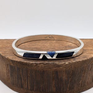 Peut inclure: Un bracelet jonc en argent avec une finition métallique polie et des segments géométriques incrustés de pierre bleu foncé et noire. L'intérieur de l'anneau porte l'inscription 925, indiquant la teneur en argent de ce bijou.