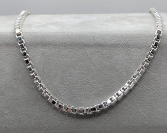 Op de afbeelding: Een zilveren box chain ketting. De ketting is gemaakt van kleine, rechthoekige schakels die met elkaar verbonden zijn. De ketting wordt gepresenteerd op een grijze ondergrond. De ketting is helder en glanzend zilver van kleur.