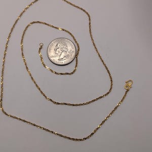 May include: A gold-colored necklace with a twisted rope design. A U.S. quarter is next to the necklace for size comparison. The necklace has a small clasp at the end.