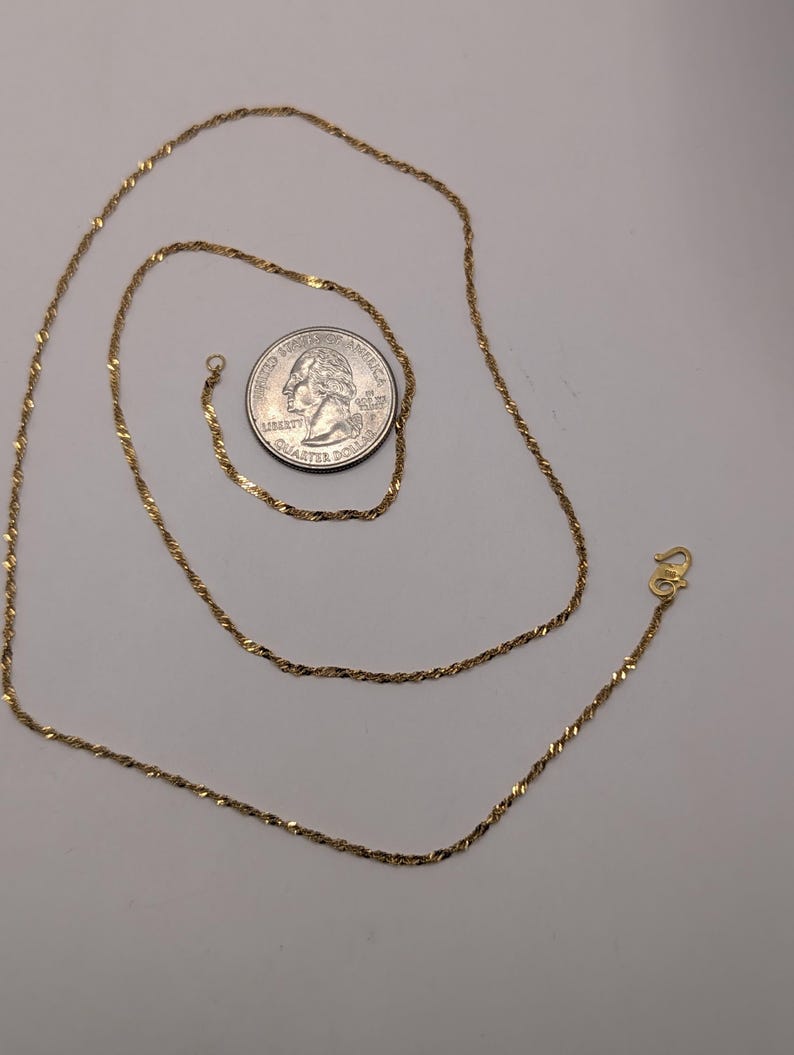 May include: A gold-colored necklace with a twisted rope design. A U.S. quarter is next to the necklace for size comparison. The necklace has a small clasp at the end.