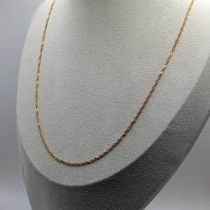 Puede incluir: Un delicado collar dorado exhibido en un maniquí. La cadena tiene un diseño de cuerda trenzada, creando una textura sutil. El collar está elegantemente drapeado, mostrando su longitud y estilo. El fondo es de color neutro, resaltando la joyería.