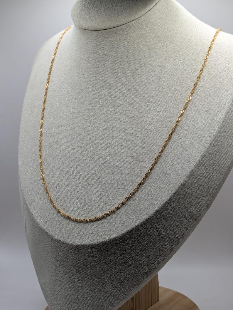 May include: A delicate gold-colored necklace displayed on a mannequin. The chain has a twisted rope design, creating a subtle texture. The necklace is draped gracefully, showcasing its length and style. The background is a neutral color, highlighting the jewelry.