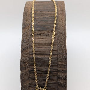 May include: A delicate gold-colored necklace with a twisted rope design. The necklace is displayed on a dark wooden stand. The clasp is visible at the bottom of the necklace. The necklace is approximately 18 inches long.