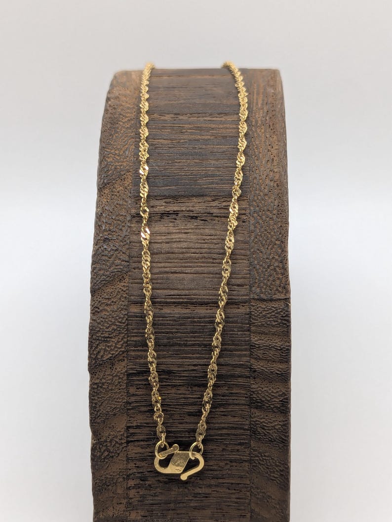 May include: A delicate gold-colored necklace with a twisted rope design. The necklace is displayed on a dark wooden stand. The clasp is visible at the bottom of the necklace. The necklace is approximately 18 inches long.