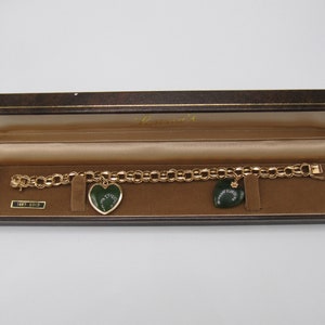 May include: A gold chain bracelet with two heart-shaped charms. The charms are green with gold accents and have a design of a clover inside. The bracelet is in a brown box with the word "Lenox" on it.