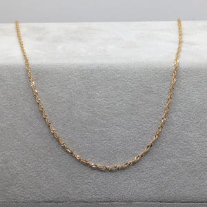 May include: A delicate gold-colored necklace with a twisted rope design. The chain is displayed on a gray surface, showcasing its subtle sparkle. This piece of jewelry is suitable for everyday wear or special occasions.