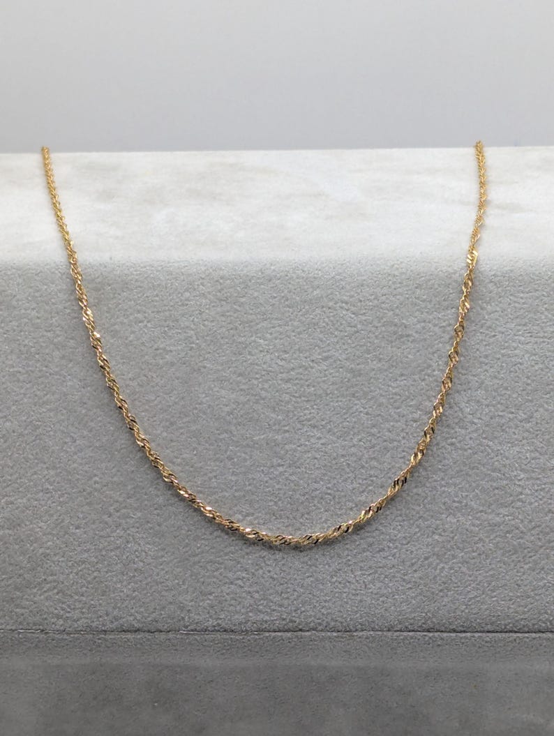 May include: A delicate gold-colored necklace with a twisted rope design. The chain is displayed on a gray surface, showcasing its subtle sparkle. This piece of jewelry is suitable for everyday wear or special occasions.